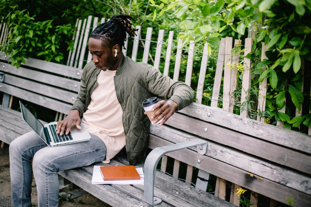 Young focused African American male student with coffee to go and notebooks working on laptop and listening to music through wireless earphones while sitting on wooden bench in green park